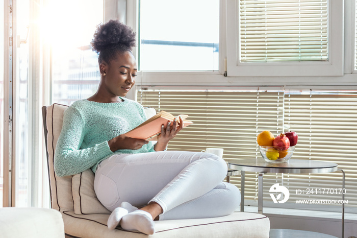 Cozy sofa and a beautiful girl, reading a book, concepts of home and comfort, place for text. Beautiful young black woman reading book near window at home. Stay home