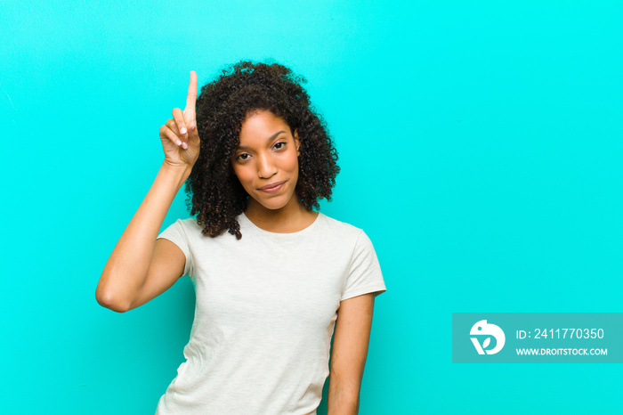 young black woman smiling cheerfully and happily, pointing upwards with one hand to copy space against blue wall