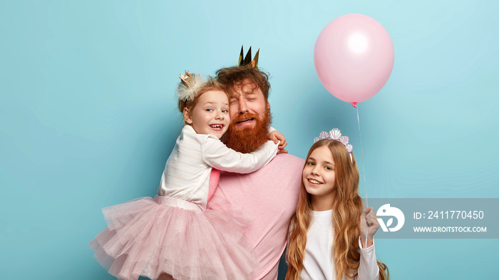 Childrens day concept. Depressed red haired father spends all day with two small daughters, feels tired after having party. Children play with balloons. Carefree childhood. Fatherhood, celebration