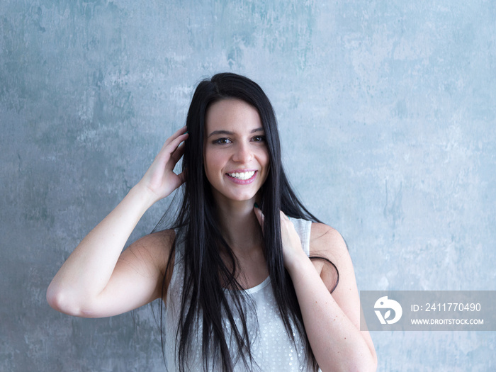 Medium horizontal portrait of beautiful smiling young woman in white sleeveless top touching her long black hair