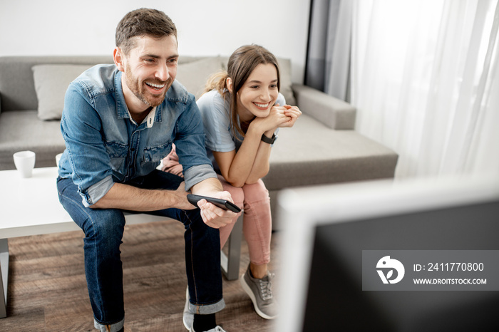 Young lovely couple with happy emotions watching TV sitting together on the table at home