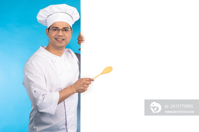 Restaurant cook. Man in chef uniform. Chef of restaurant shows white banner. Guy confectioner smiles standing on blue. Place for inscription on white. Happy chef. Successful restaurant owner