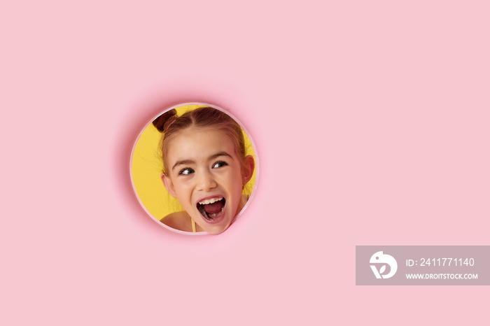 super excited little child girl looking aside through the hole on pink background