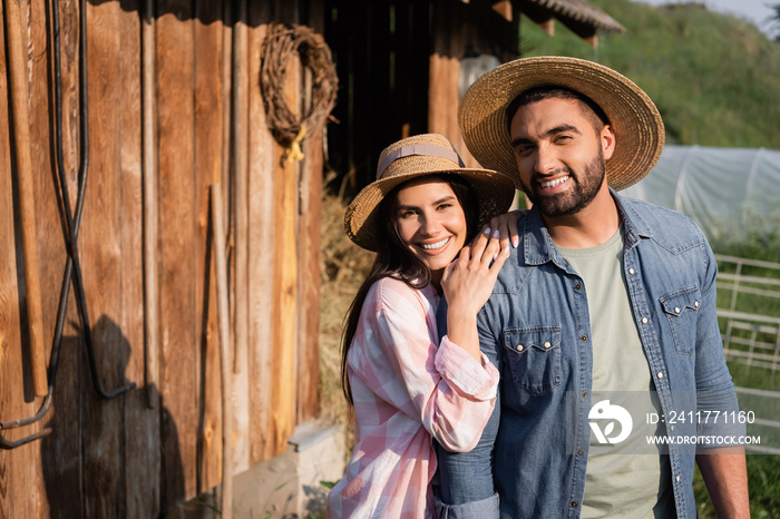 pleased farmers in straw hats looking at camera near wooden barn on summer day.