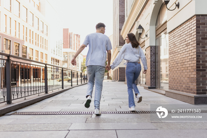 Full-length portrait from behind of young happy couple walking around city on summer sunny day