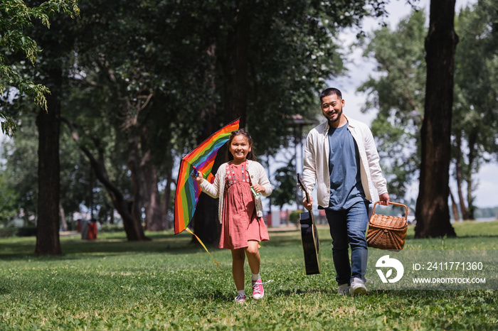 Positive asian family with flying kite and acoustic guitar in summer park.