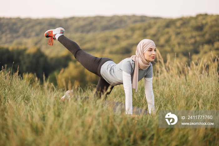 Smiling young woman in hijab and sport outfit doing flexible exercises on yoga mat during morning workout on fresh air. Training at summer park. Healthy lifestyles.