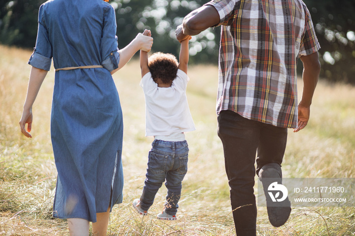 Back view of mixed race parents walking with little son on summer field and holding hands. Close up of happy family of three playing together outdoors. Love and happiness concept.