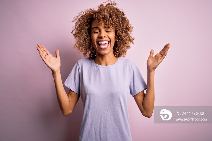 Young beautiful african american woman wearing casual t-shirt standing over pink background celebrating mad and crazy for success with arms raised and closed eyes screaming excited. Winner concept