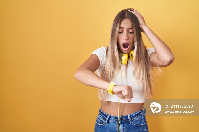 Young blonde woman standing over yellow background wearing headphones looking at the watch time worried, afraid of getting late