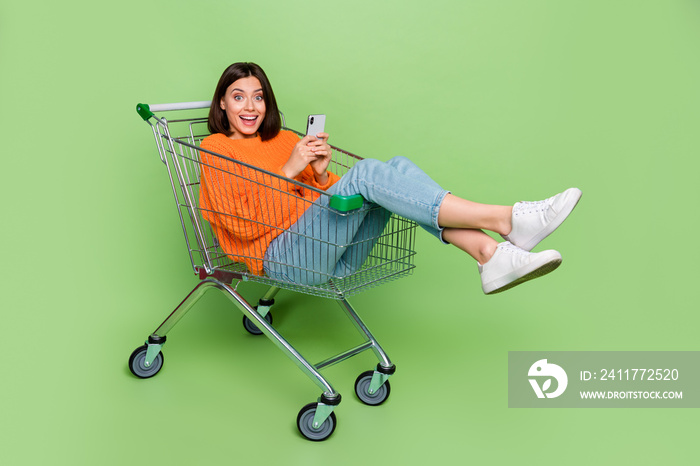 Portrait of attractive trendy cheerful girl riding cart using device smm blogging isolated over bright green color background