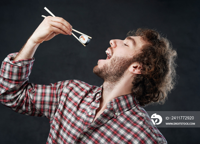 Young stylish man with curly hairstyle wearing checkered shirt eating sushi rolls on the dark background