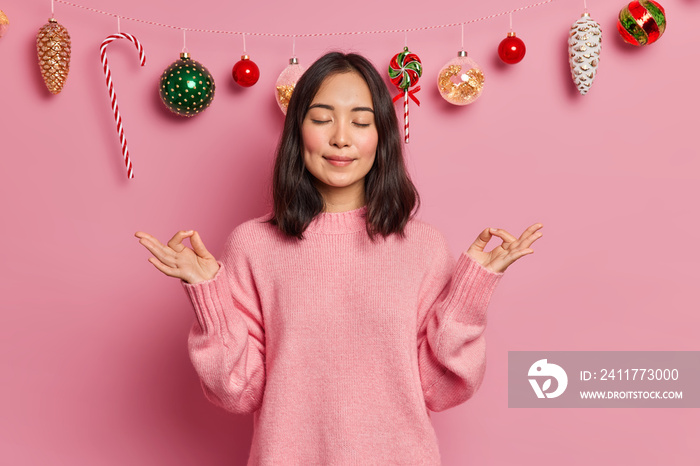 Photo of relaxed brunette woman in casual sweater does meditation gesture with fingers practices yoga on New Year Eve closes eyes has calm expression stands against garland made on christmas toys