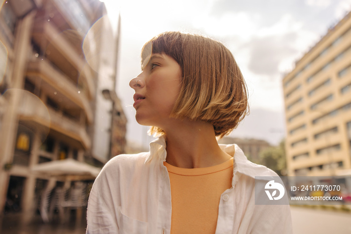 Close-up of pretty young caucasian woman looking away standing on street. Girl with brown short hair wears t-shirt and shirt. Lifestyle concept