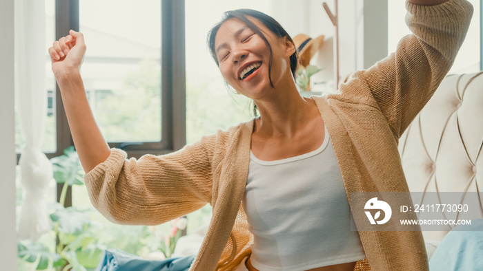 Asian girl wear white cream pajamas cardigan on fluffy comfort bed happy dancing with cheerful moment for long holiday in bedroom at home in morning light from window. Female morning vibes concept.