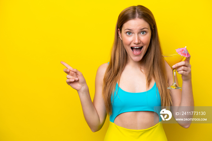 Young caucasian woman holding a cocktail isolated on yellow background surprised and pointing finger to the side