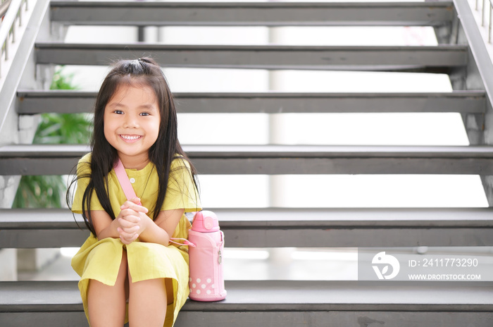 Asian child cute or kid girl smiling happy and sling water bottle or flask and sit on stairs for relax on holiday travel or waiting study to summer and thinking for new idea at nursery pre school