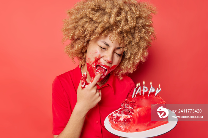 Beautiful curly haired woman licks finger eats sweet creamy cake with hands has spoiled makeup poses indoor against vivid red background. Female model enjoys appetizing dessert during holiday