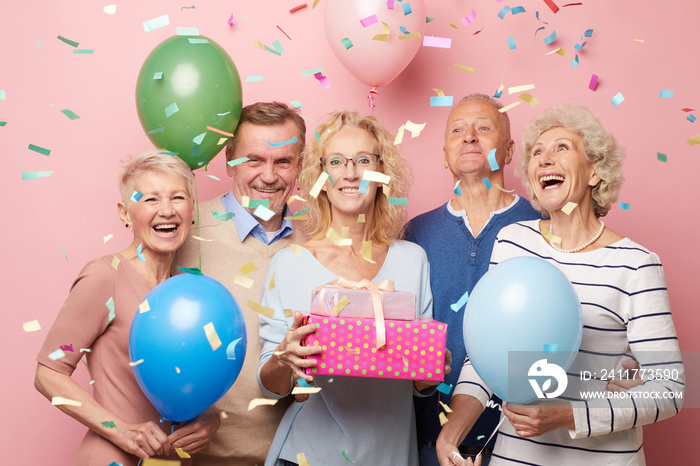 Cheerful excited mature friends in casual outfits standing against pink wall under falling confetti and having fun together at birthday party