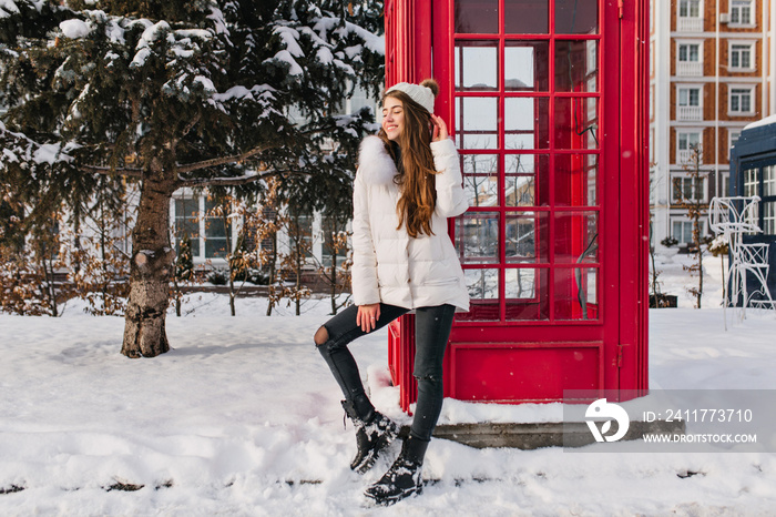 Full-length portrait of enthusiastic lady with long hairstyle posing near red call-box in winter. Outdoor photo of pretty caucasian woman in white hat enjoying december vacation in England..