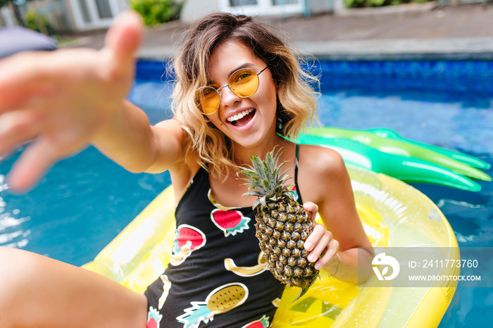 Amazing short-haired girl in trendy glasses swimming in pool. Outdoor photo of lovely tanned female model in stylish swimsuit laughing in summer weekend.