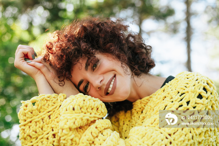 Close-up outdoor portrait of attractive young woman with curly hair and smiling. Female has positive expression, wearing knitted yellow sweater and posing against nature background. People, lifestyle