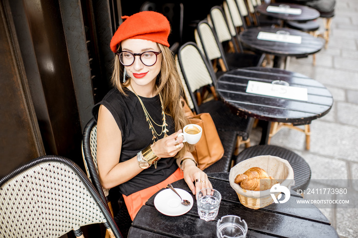 Young stylish woman in red beret having a french breakfast with coffee and croissant sitting oudoors at the cafe terrace