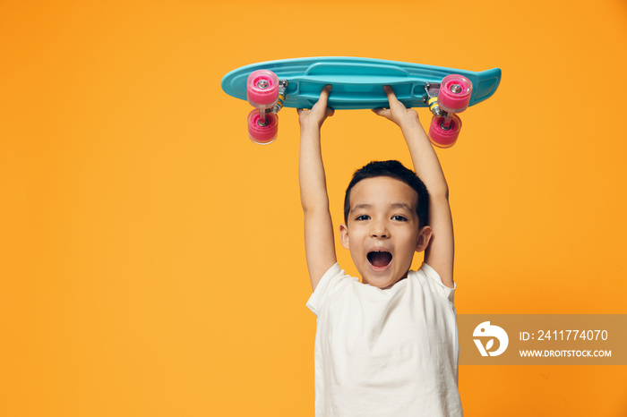 happy, funny little boy stands smiling broadly against an orange background raising his blue skateboard high in his hands