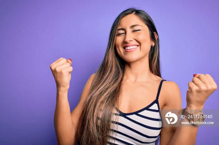 Young beautiful woman wearing casual summer striped top over purple isolated background very happy and excited doing winner gesture with arms raised, smiling and screaming for success. Celebration