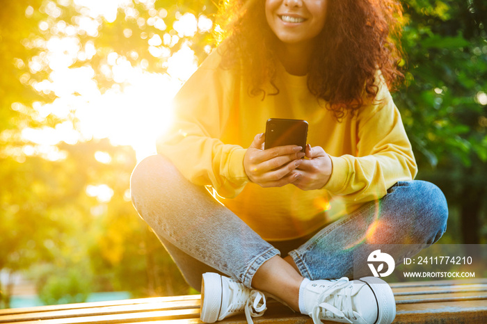 Cropped image of charming young woman smiling and holding smartphone while sitting on bench in green park