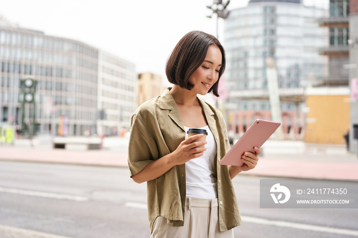 Happy city girl walking on streets, using tablet and drinking coffee on her way to work