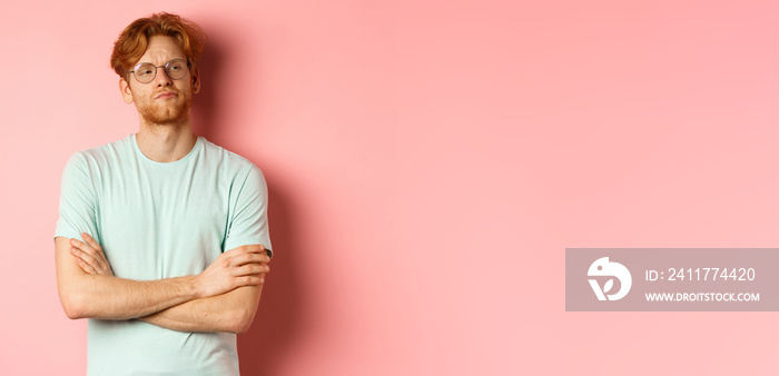 Arrogant redhead guy in glasses cross arms on chest, looking at something with skeptical face, standing over pink background