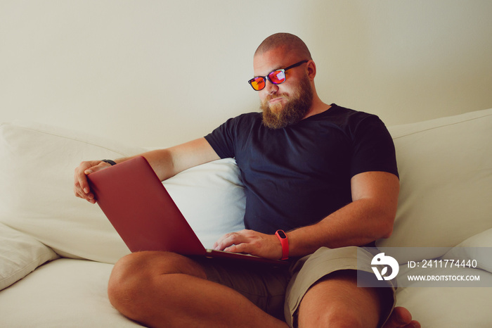 Bearded Man with blue light blocking eye glasses (yellow amber lens) working with laptop sitting on sofa