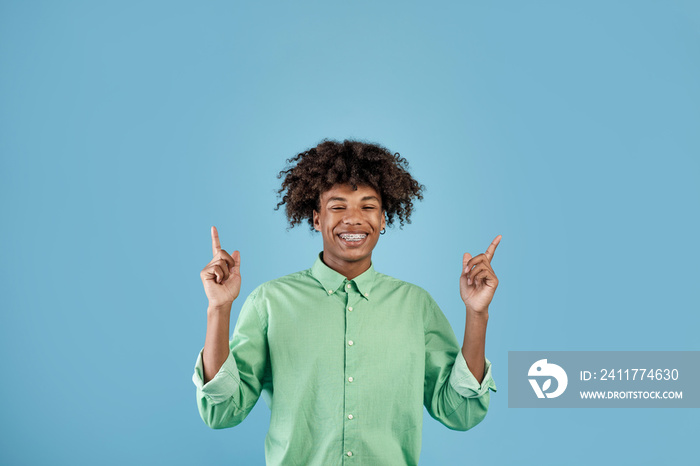 Look up. Happy african american guy pointing fingers upward and smiling at camera, showing something, blue background