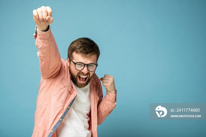 Joyful, smiling bearded man raises his hand up, feeling victory and success. Copy space. Isolated on a blue background.