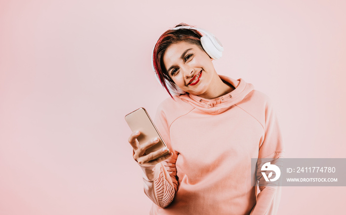 young hispanic woman girl listening music with headphones  and holding mobile phone on coral pink background with copy space studio portrait in Mexico Latin America