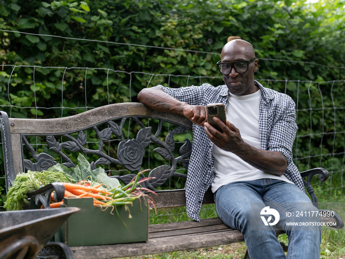 Smiling mature man resting on bench and using phone after working in garden