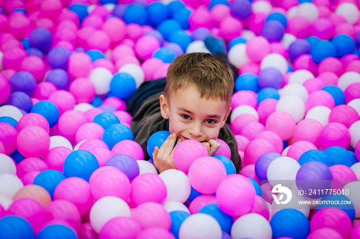 A beautiful little smiling boy, a preschool child lies in multi-colored plastic balls on the playground. Photography, portrait, childhood vacation concept.