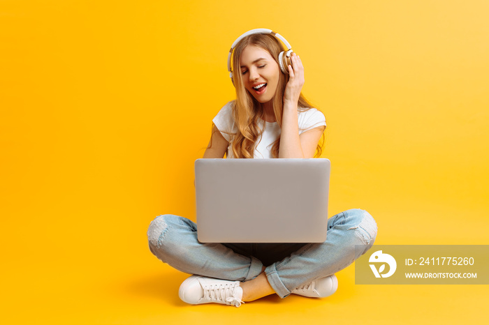 An image of a beautiful woman listening to music on headphones using a laptop while sitting cross-legged on a yellow background