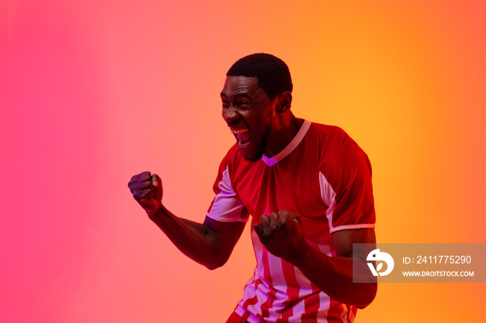 Happy african american male soccer player raising hands over neon pink lighting