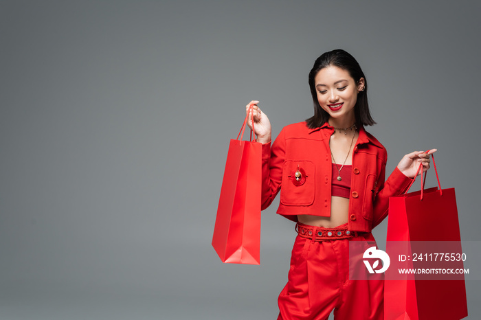 happy asian woman in elegant clothes posing with red shopping bags isolated on grey.