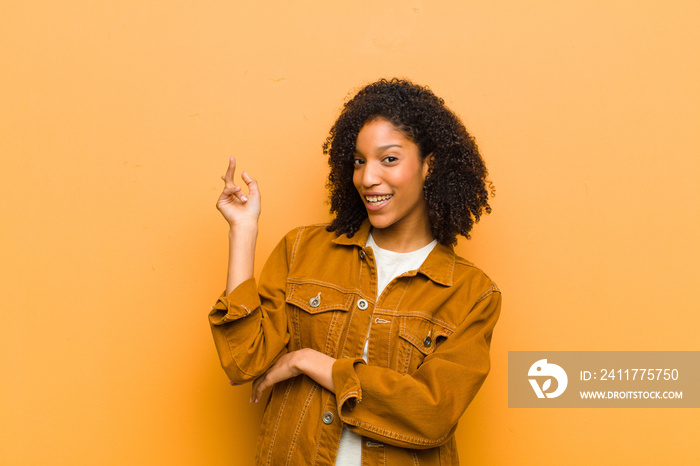 young pretty black woman smiling happily and looking sideways, wondering, thinking or having an idea against orange wall