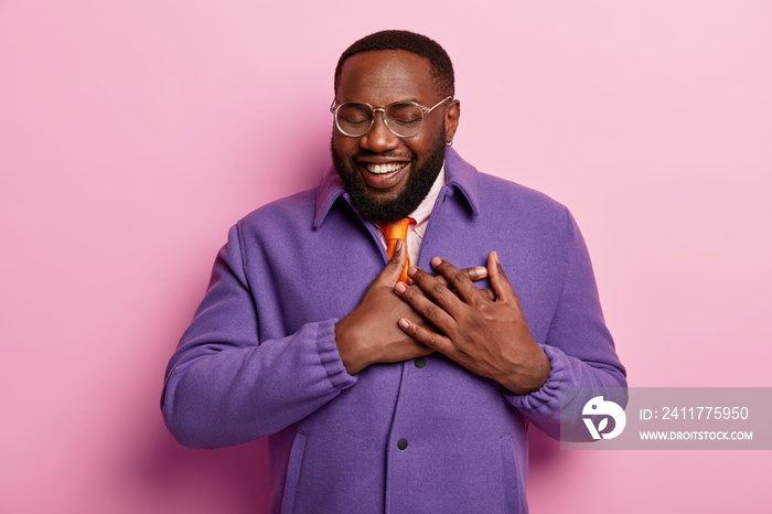 Touched happy bearded black man hears heartwarming words, expresses kindness, wears transparent glasses, wears optical glasses, purple jacket, good feelings, isolated over pastel rosy background