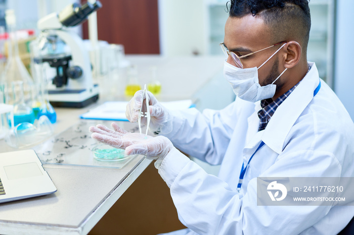 Serious experienced young Arabian male scientist using tweezers and putting blue crystal on hand in glove while working in medical laboratory