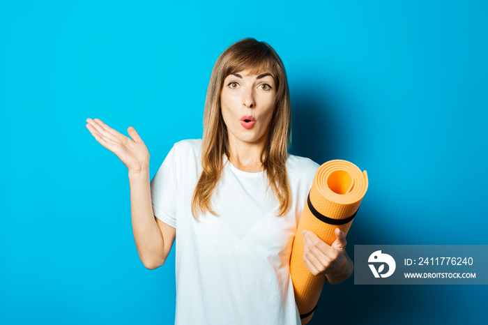 Young woman in a white T-shirt with a surprised face holds a yoga mat and makes a hand gesture on a blue background. Concept of yoga, yoga for beginners, fitness, stretching