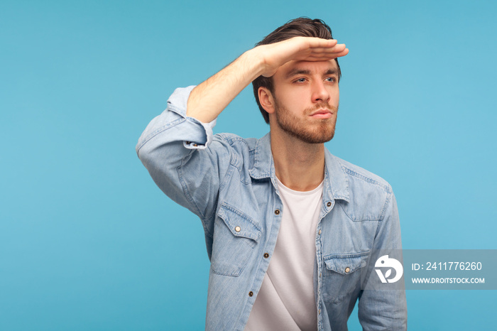 Far future. Portrait of handsome man in worker denim shirt looking into distance, holding hand over eyes and watching horizon with attentive view. indoor studio shot isolated on blue background