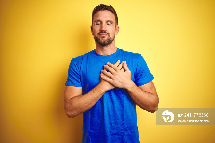 Young handsome man wearing casual blue t-shirt over yellow isolated background smiling with hands on chest with closed eyes and grateful gesture on face. Health concept.