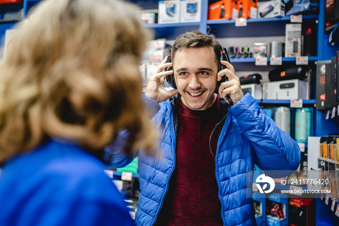 Front view portrait of young adult caucasian man standing by female seller in the electronics store looking and peaking products checking earphones wearing blue jacket real people copy space