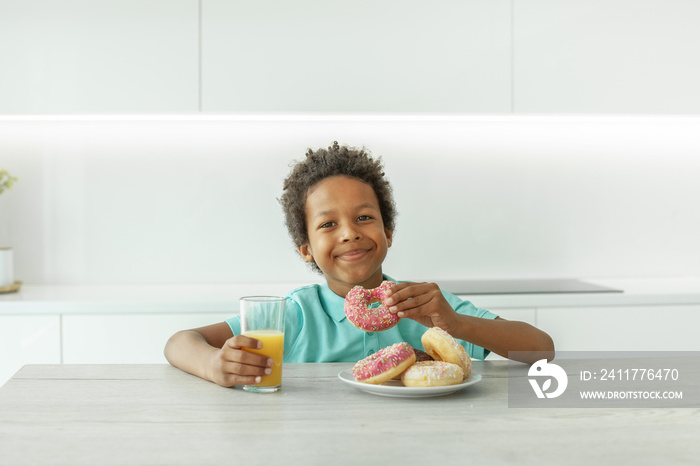 Smiling child boy drinking orange juice and eating donuts in white kitchen