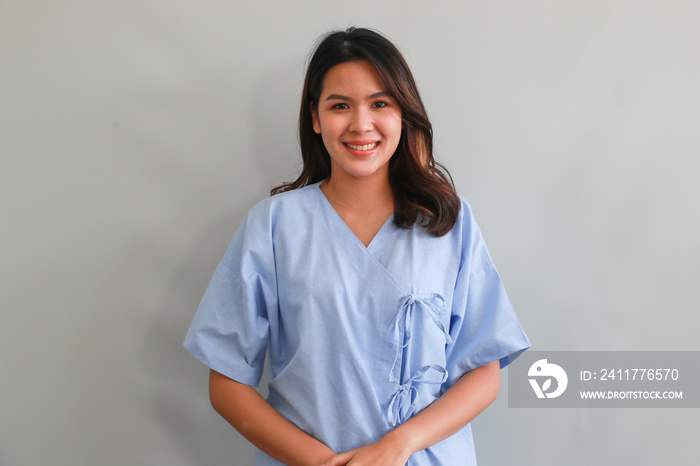 Young Asian female patient in blue hospital patient gown smiling and looking at camera on grey background, authentic half body portrait. Medical and healthcare concept.